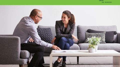 Two people seated on a gray sofa, talking and reviewing documents on a white coffee table, with a green banner and award logo above.